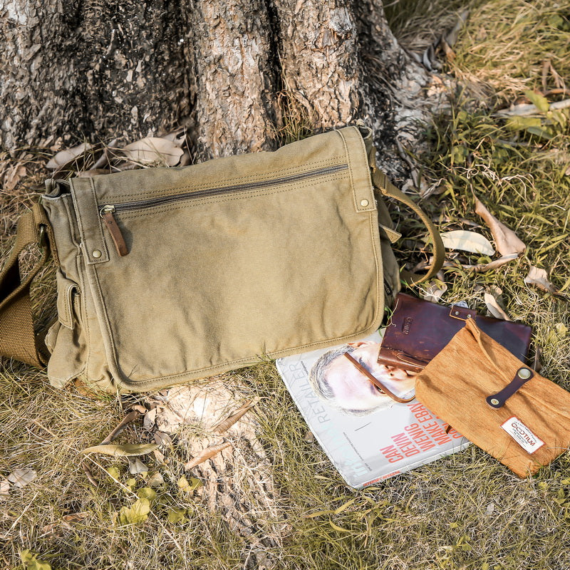 An olive green canvas messenger bag with brown leather accents, styled outdoors on the ground next to a tree trunk with a magazine and leather accessories.