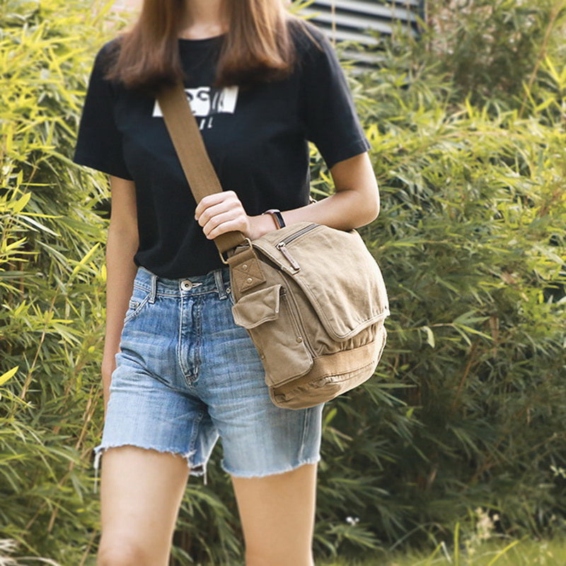 A woman wearing a black graphic t-shirt and denim shorts carries a beige canvas messenger bag, set against a background of green bamboo foliage.