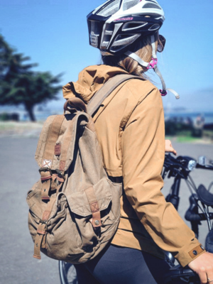A person wearing a bicycle helmet and tan jacket, seen from behind while riding a bicycle outdoors. They are carrying a large brown canvas backpack with multiple front pockets and brown leather-like straps.
