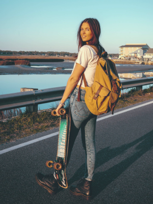 A woman stands on a road beside water at sunset, holding a skateboard and wearing a tan backpack, white T-shirt, blue jeans, and brown boots.