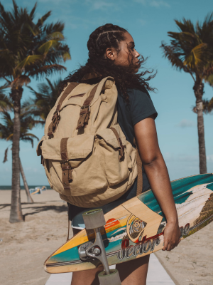 A woman on a beach wearing a large beige canvas backpack and holding a colorful skateboard, with palm trees and blue sky in the background.